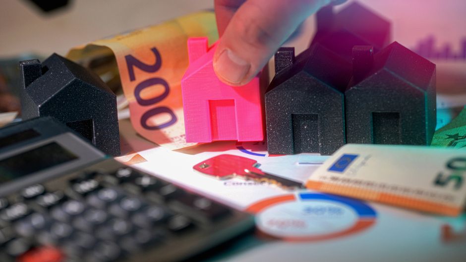 A close-up view of a hand holding a pink miniature house model, positioned among small black house models on a cluttered surface. The scene includes a rolled-up 200 euro banknote, a calculator, and va