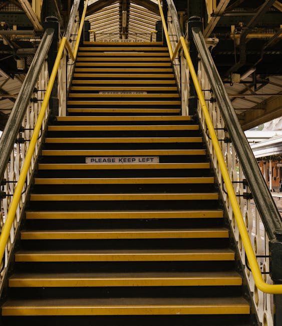 A set of metal and wooden stairs with yellow handrails leading up to an outdoor platform, likely part of a railway station or urban infrastructure near Rainham Station. The stairs have black non-slip 